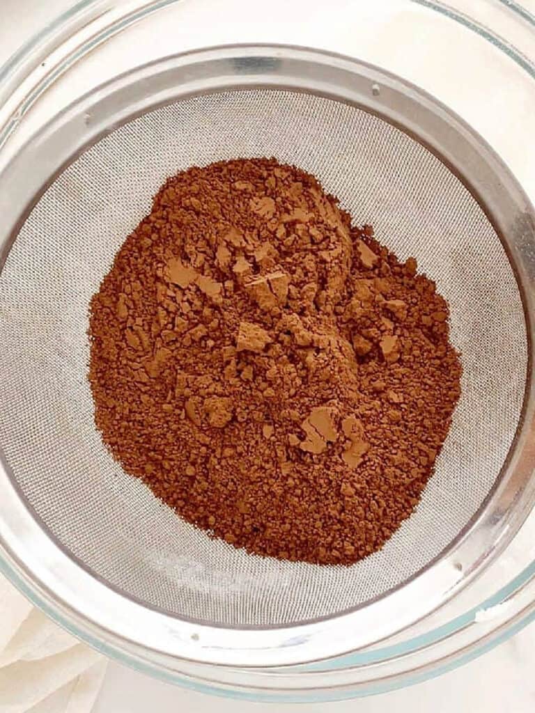 Sifting cocoa powder into a glass bowl on a white surface.
