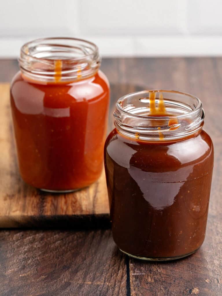 Filled jars of homemade barbecue sauce. Wooden surface, white tile background.