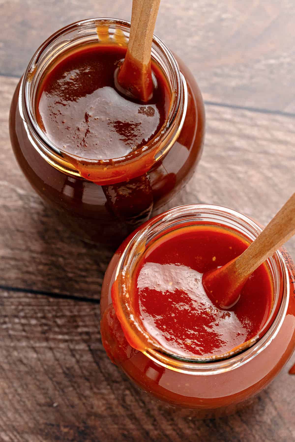Top view of two jars with barbecue sauce and wooden spoons. Wooden surface.