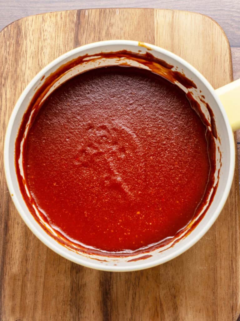 Cooked homemade BBQ sauce in a white saucepan. Wooden board surface.