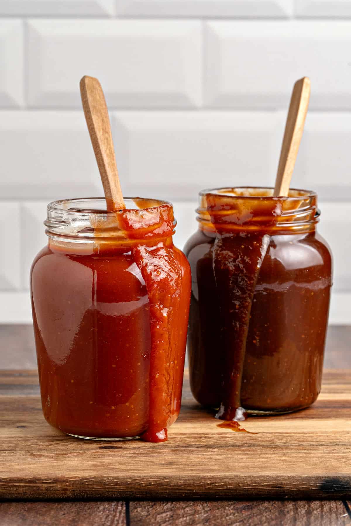 Two different barbecue sauces in jars on a wooden board. Wooden spoons, subway tile background.