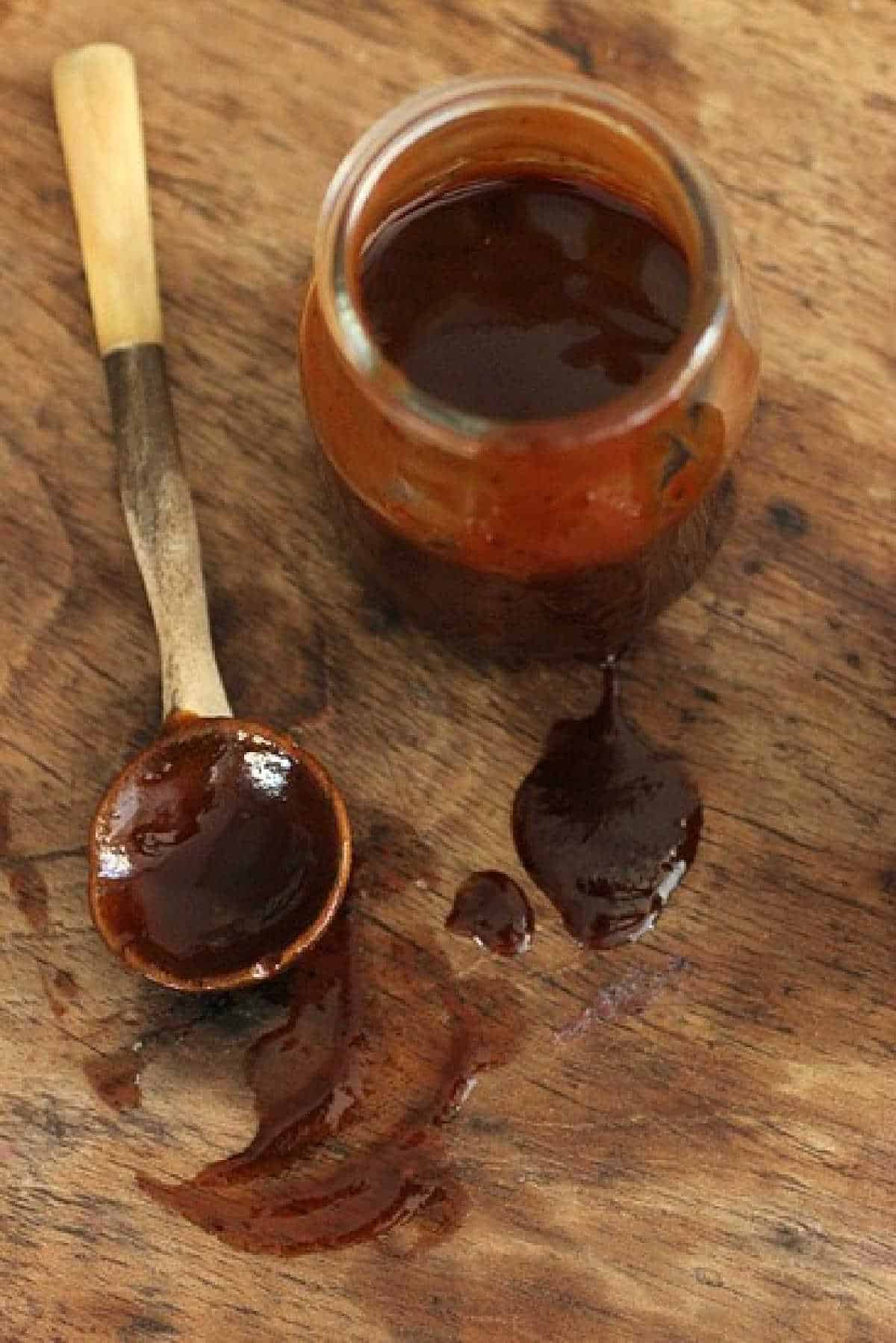 Brown barbecue sauce in jar with spoon beside it on a smeared wooden table.