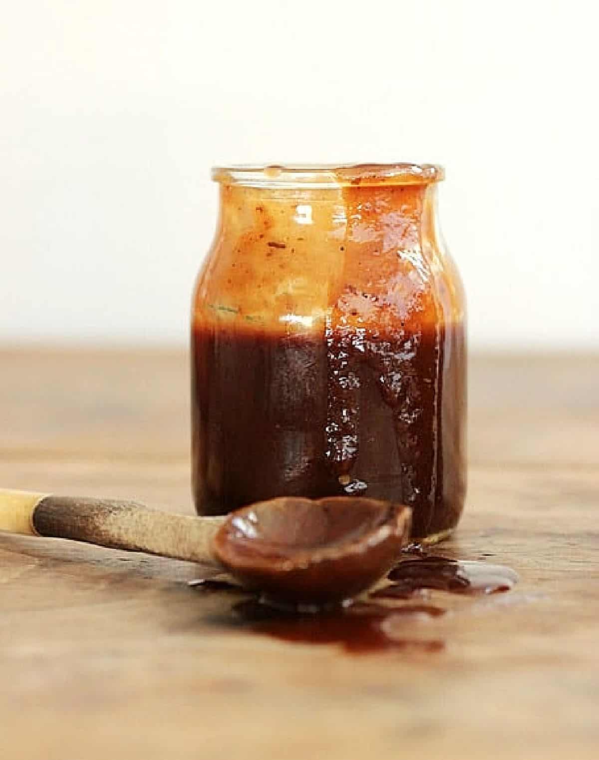 Dripping jar with brown barbecue sauce, a wooden spoon on a wooden surface.