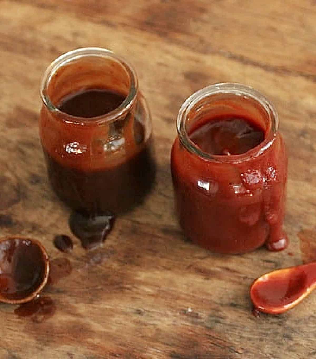 Brown and red barbecue sauces in jars on a wooden surface with spoons.