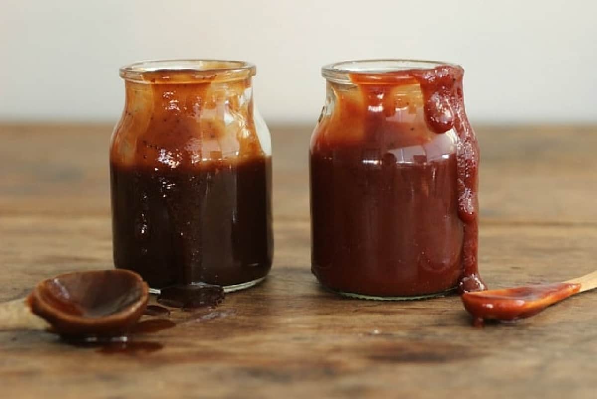 Front view of two jars with different barbecue sauces with spoons on a wooden table.