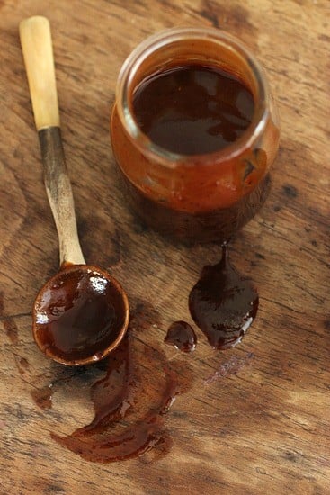 Top view of a glass jar of barbecue sauce, smeared wooden table and spoon