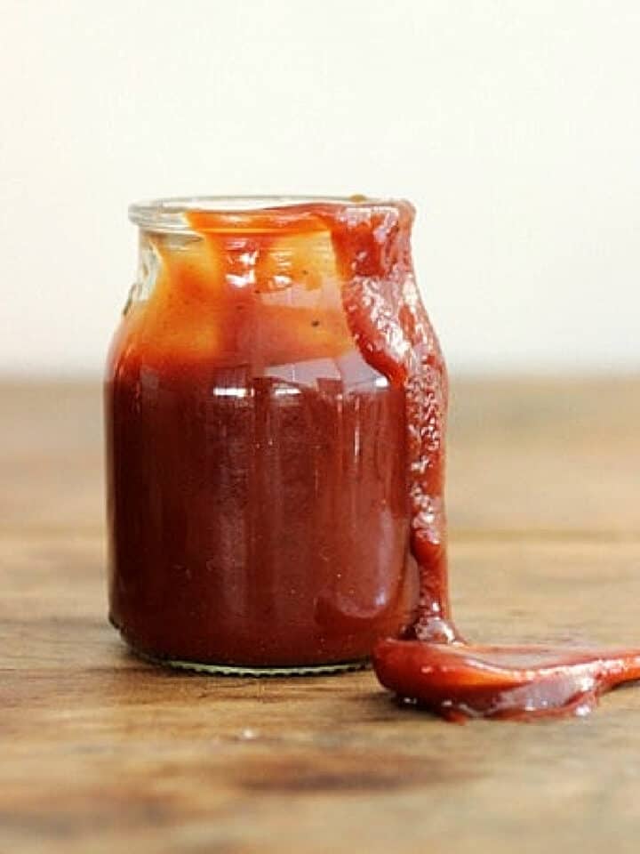 Dripping glass jar of barbecue sauce on wooden table, a spoon beside