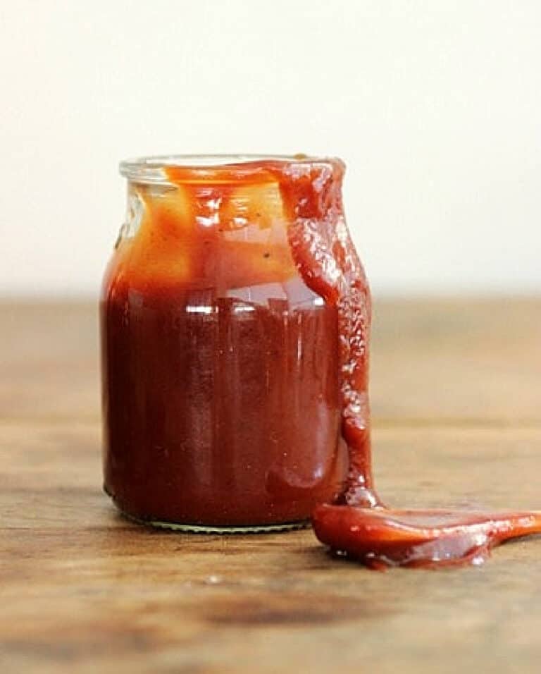Dripping glass jar of barbecue sauce on wooden table, a spoon beside