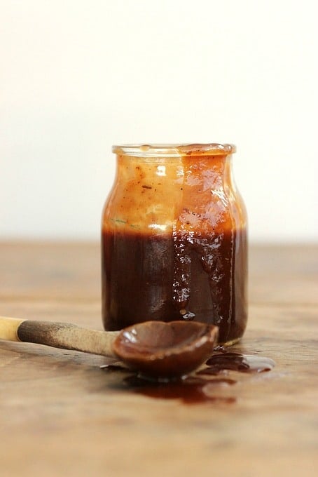 A dripping jar of brownish Barbecue Sauce on wooden table, a wooden spoon beside