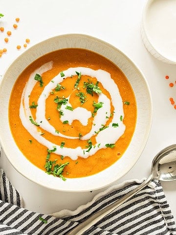 Striped blue cloth, white bowl with cream-swirled carrot lentil soup. White surface, loose red lentils.