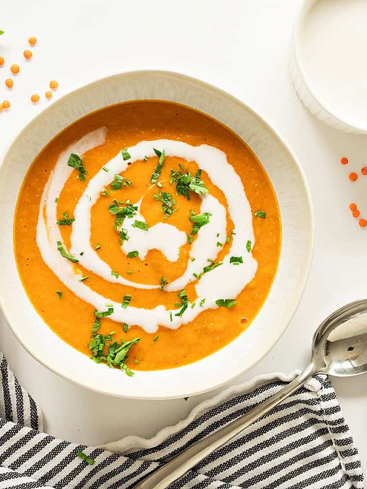 Striped blue cloth, white bowl with cream-swirled carrot lentil soup. White surface, loose red lentils.