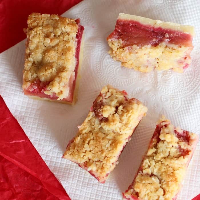 Top view of crumb bars with red filling on a white paper napkin on a red surface.