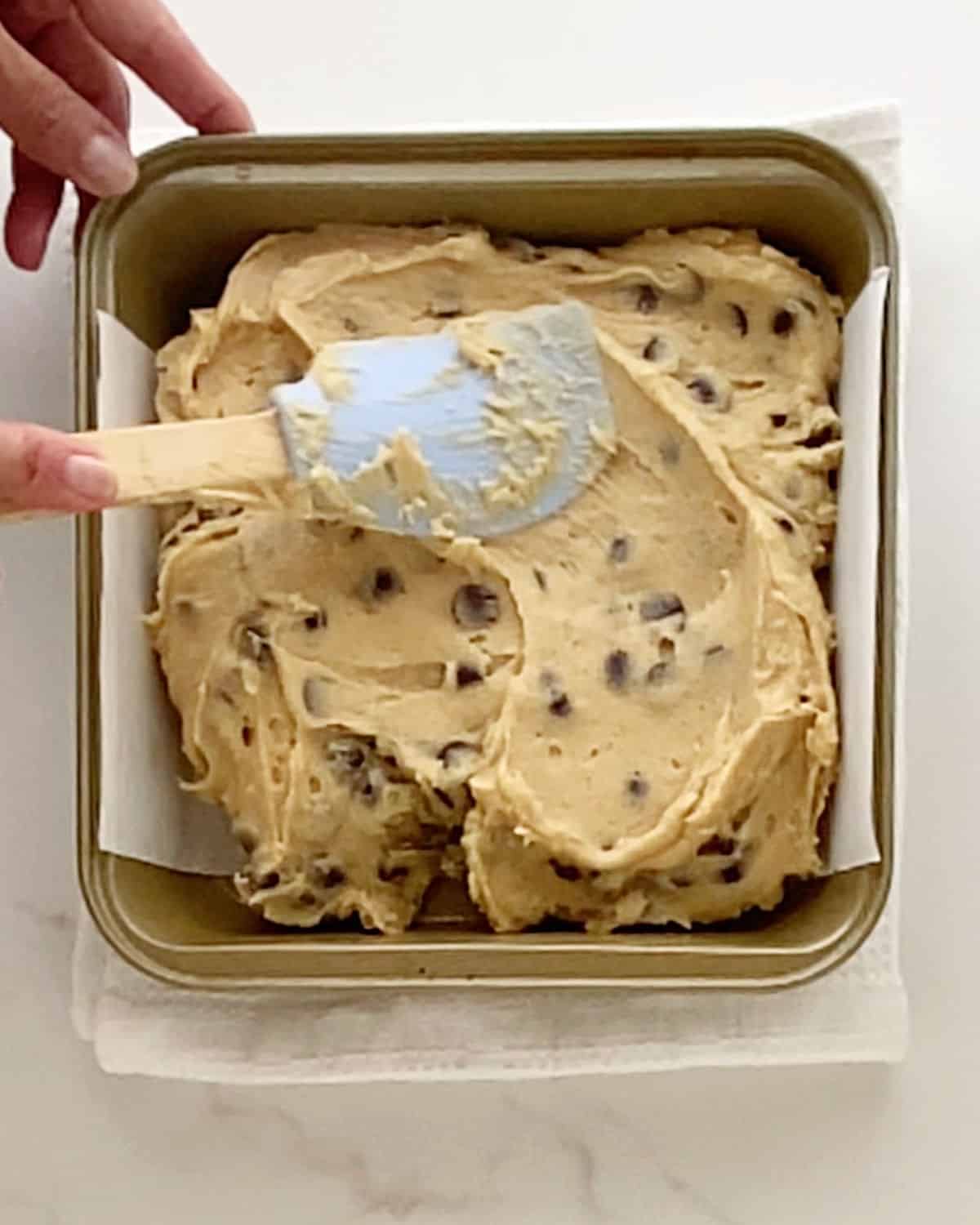 Spreading chocolate chip blondies dough in a metal pan with a spatula. White surface.