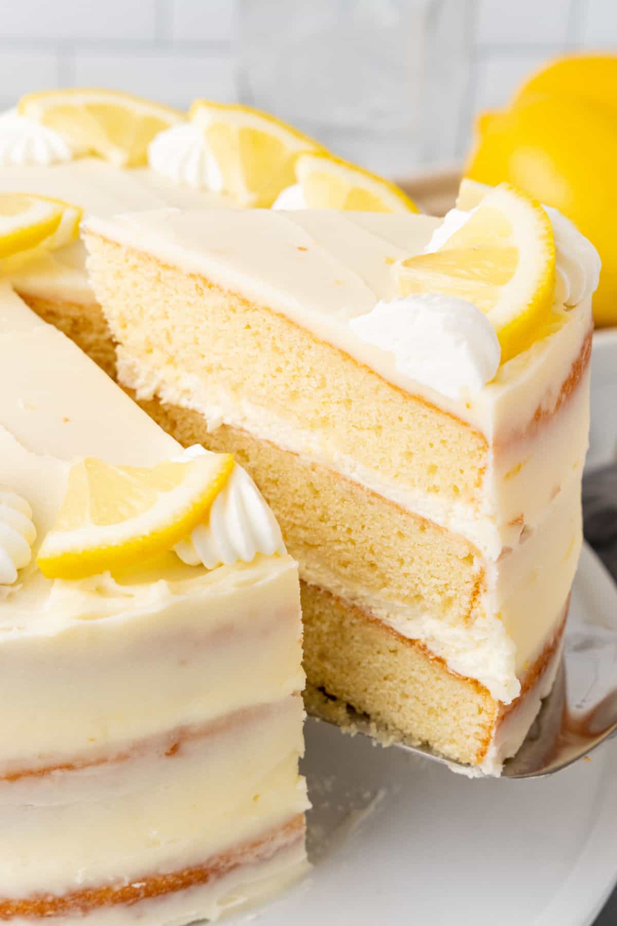 Slice of lemon layer cake being pulled from whole cake on a white plate. 