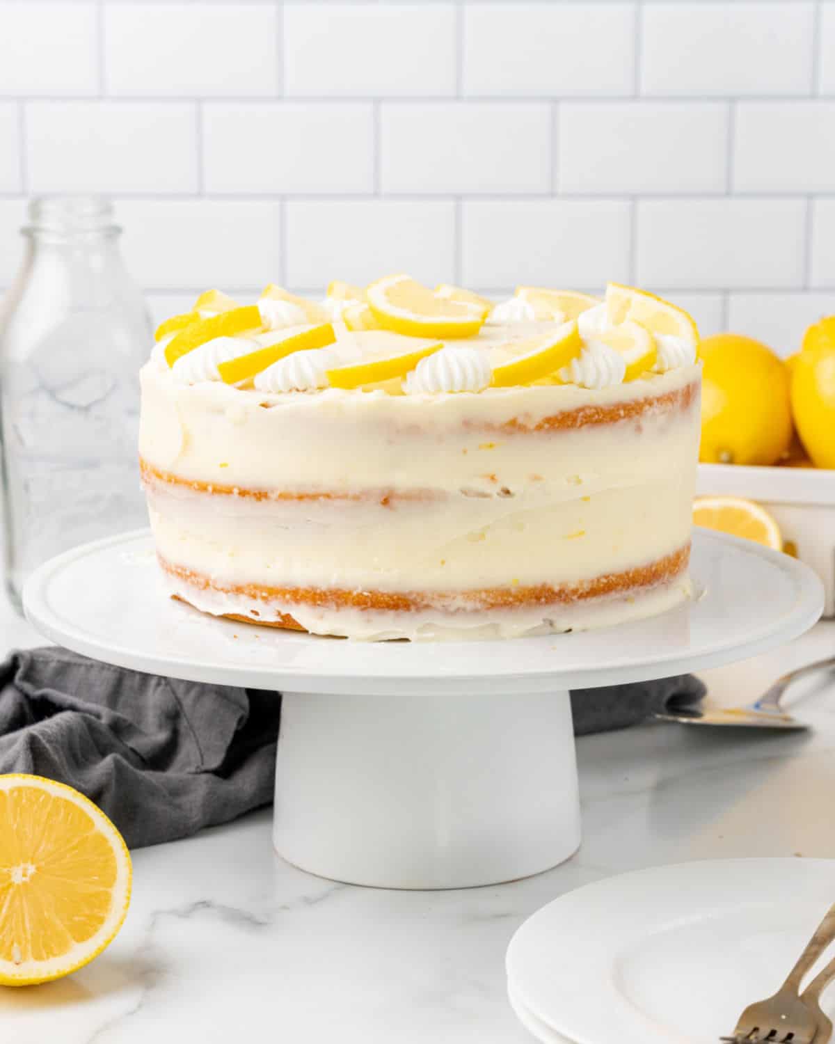 Lemon layer cake frosted and decorated on a white cake stand. White subway tiles as background.