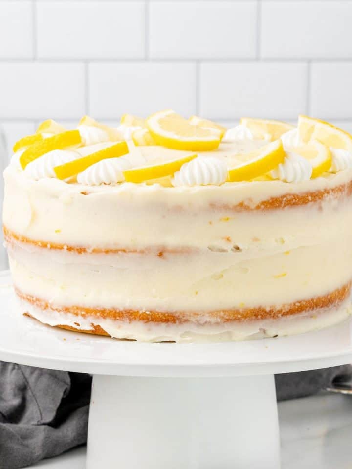 Whole frosted lemon cake on a white cake stand. Subway tiles as background.
