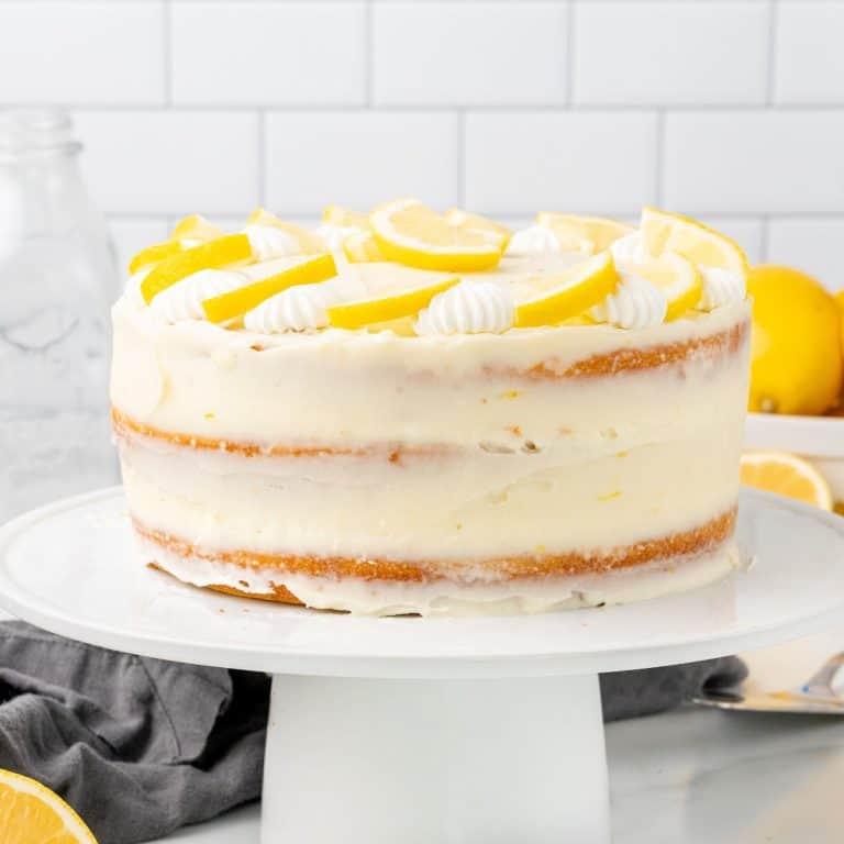 Whole frosted lemon cake on a white cake stand. Subway tiles as background.