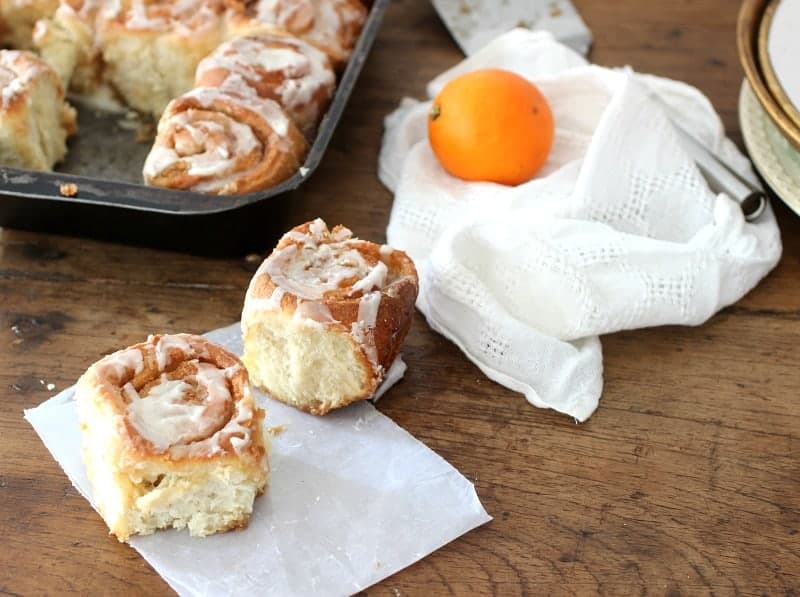 Two cinnamon rolls on wooden table, rest in pan, white linen and orange in background