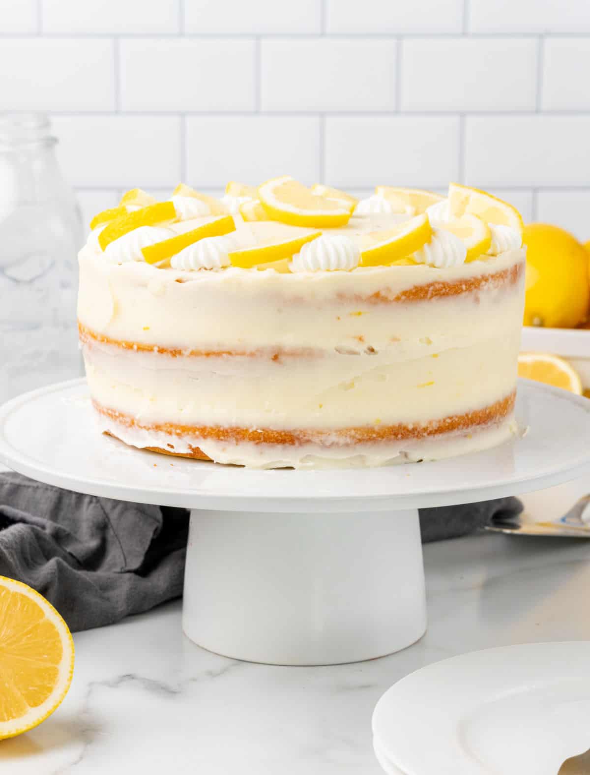 Whole frosted lemon layer cake on a white cake stand. White surface and tile background.