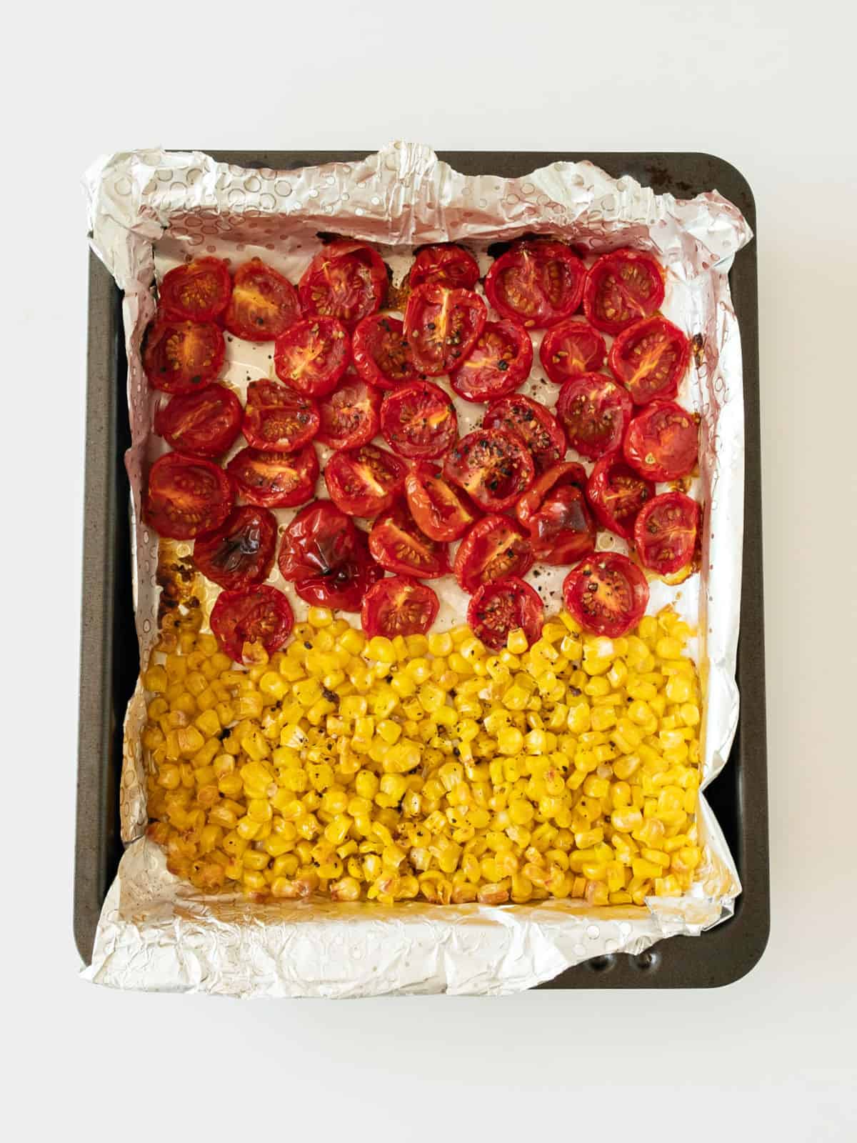 Roasted cherry tomatoes and corn on aluminum foil on a baking tray. White surface.