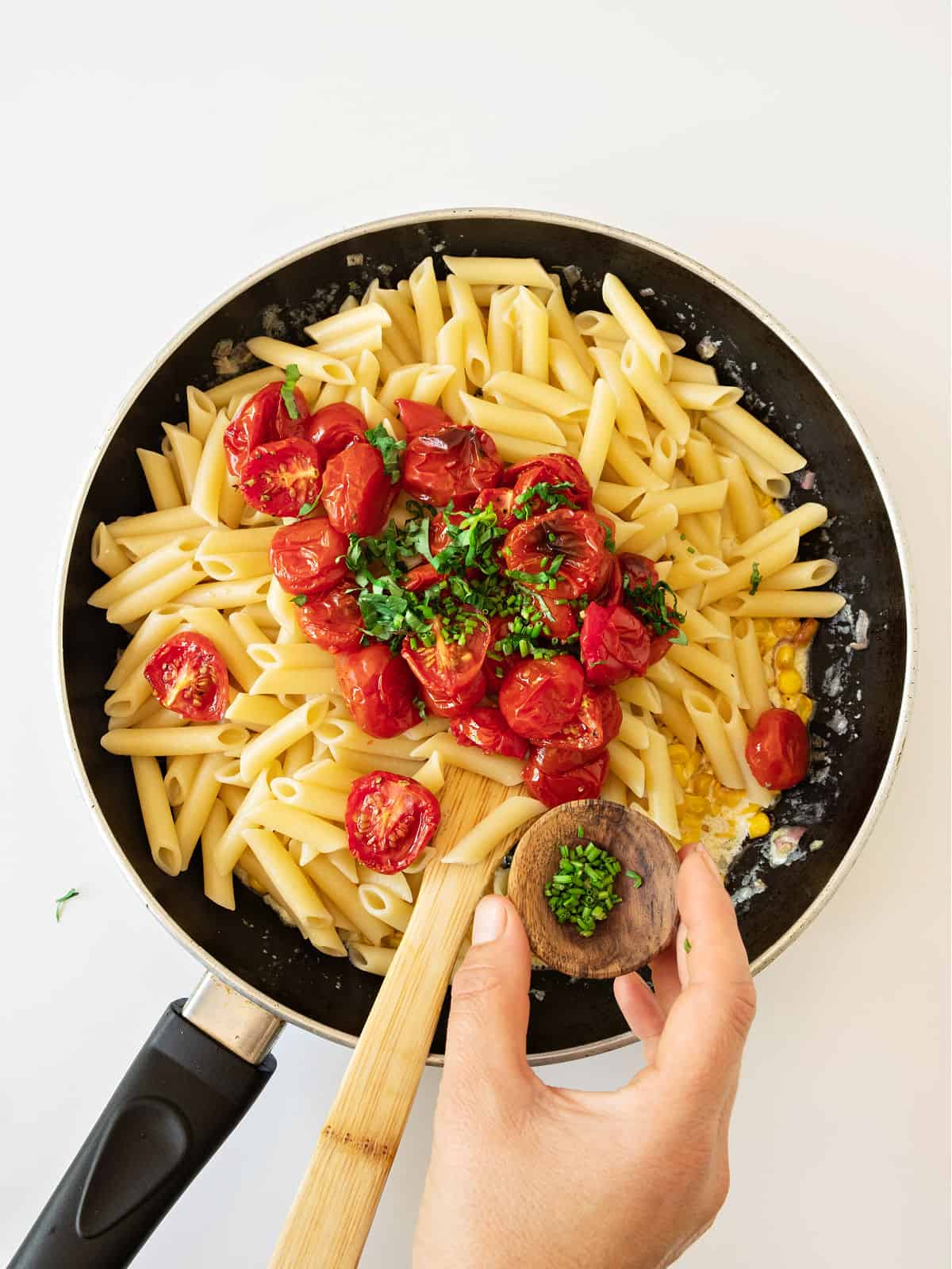Adding herbs to short pasta with tomatoes in a skillet. White surface.