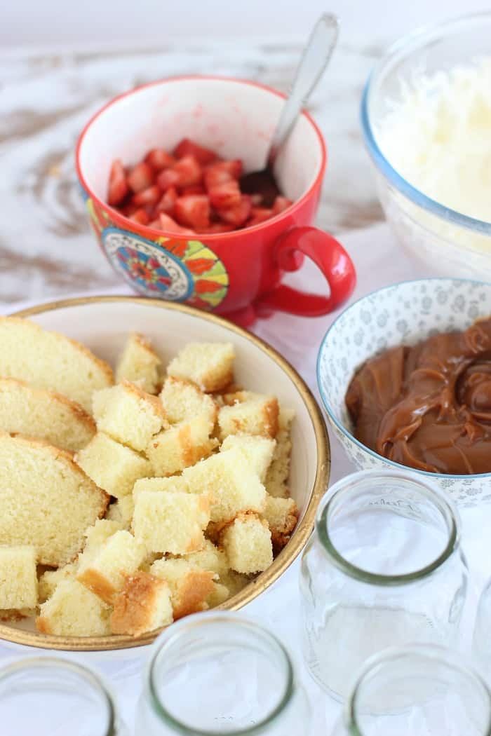 Bowls with strawberries, diced cake dulce de leche and cream, glass jars, white table