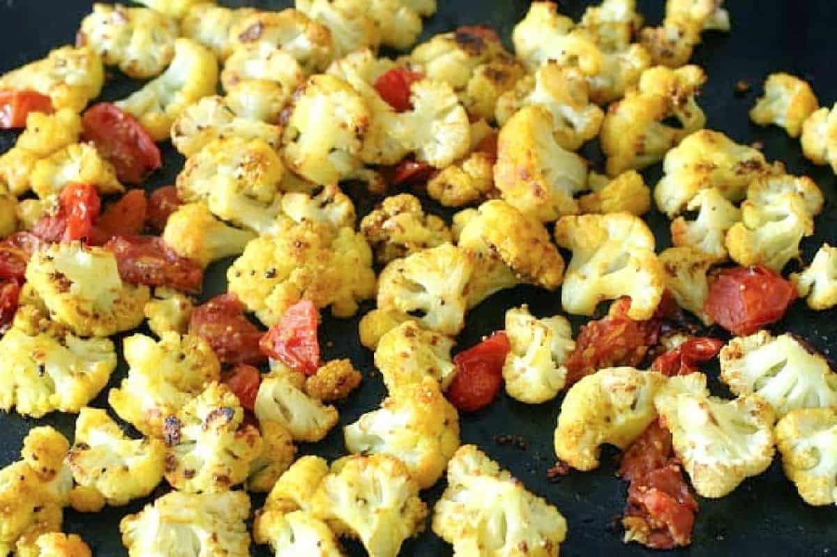 Close-up of baked cauliflower and tomatoes with turmeric on a dark baking tray.