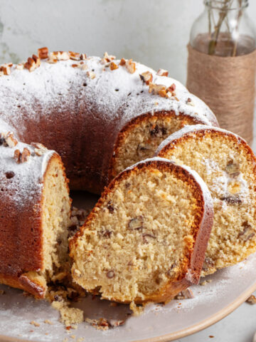 Whole and slices of coconut bundt cake with powdered sugar on a beige plate. Close up image.