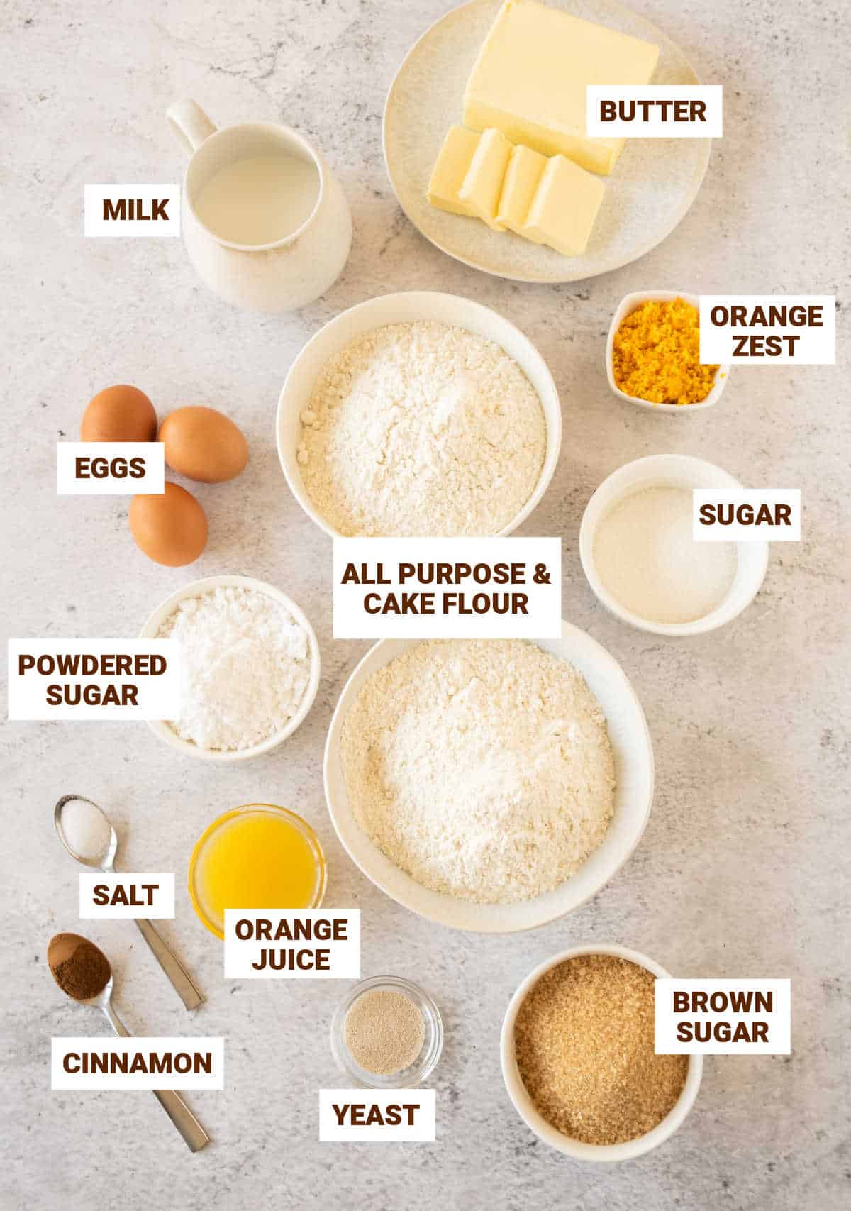 Light gray surface with bowls containing ingredients for orange cinnamon rolls including butter, flours, eggs, milk, sugar.