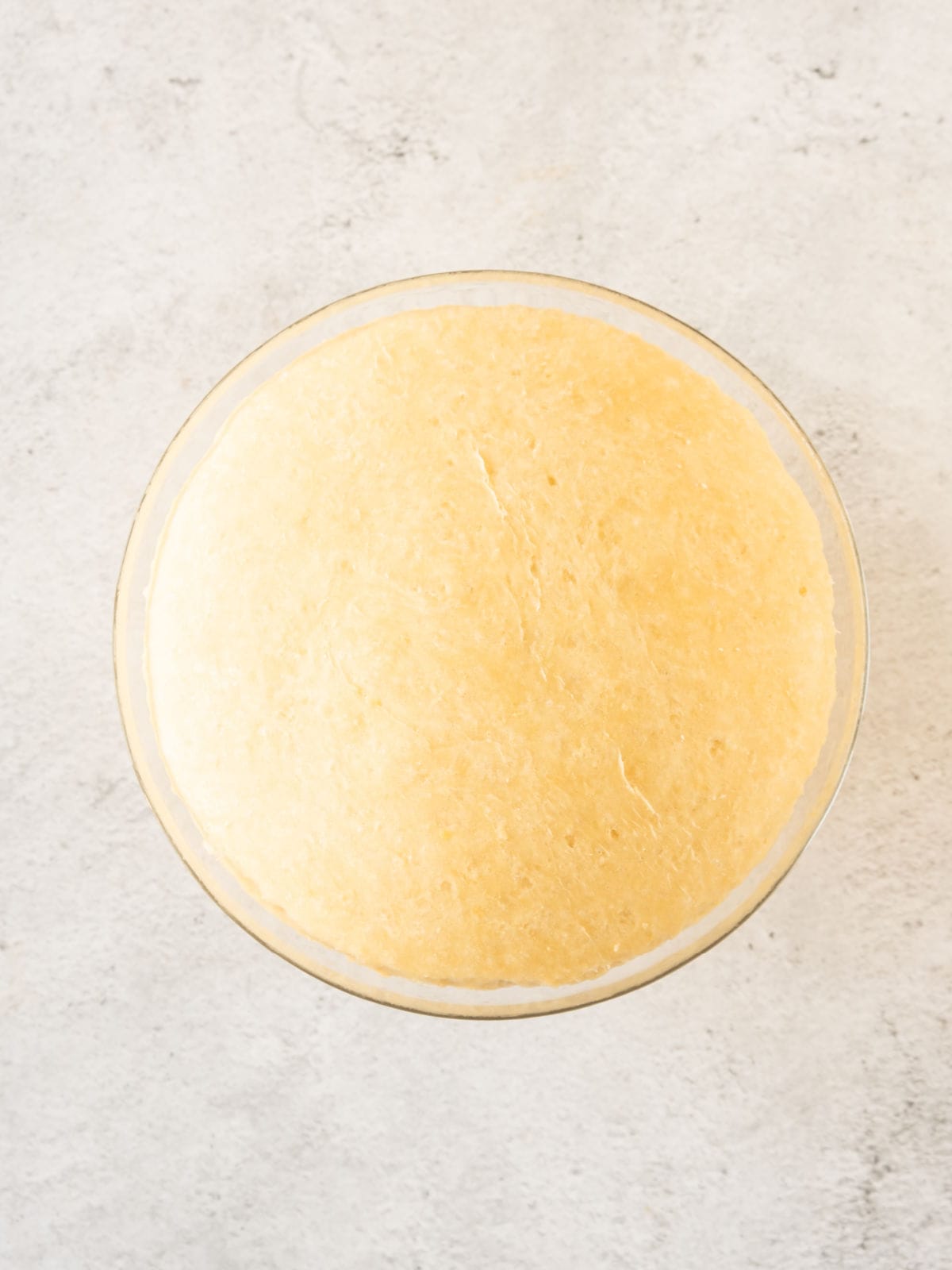 Cinnamon roll dough doubled in size in a glass bowl on a light gray surface.