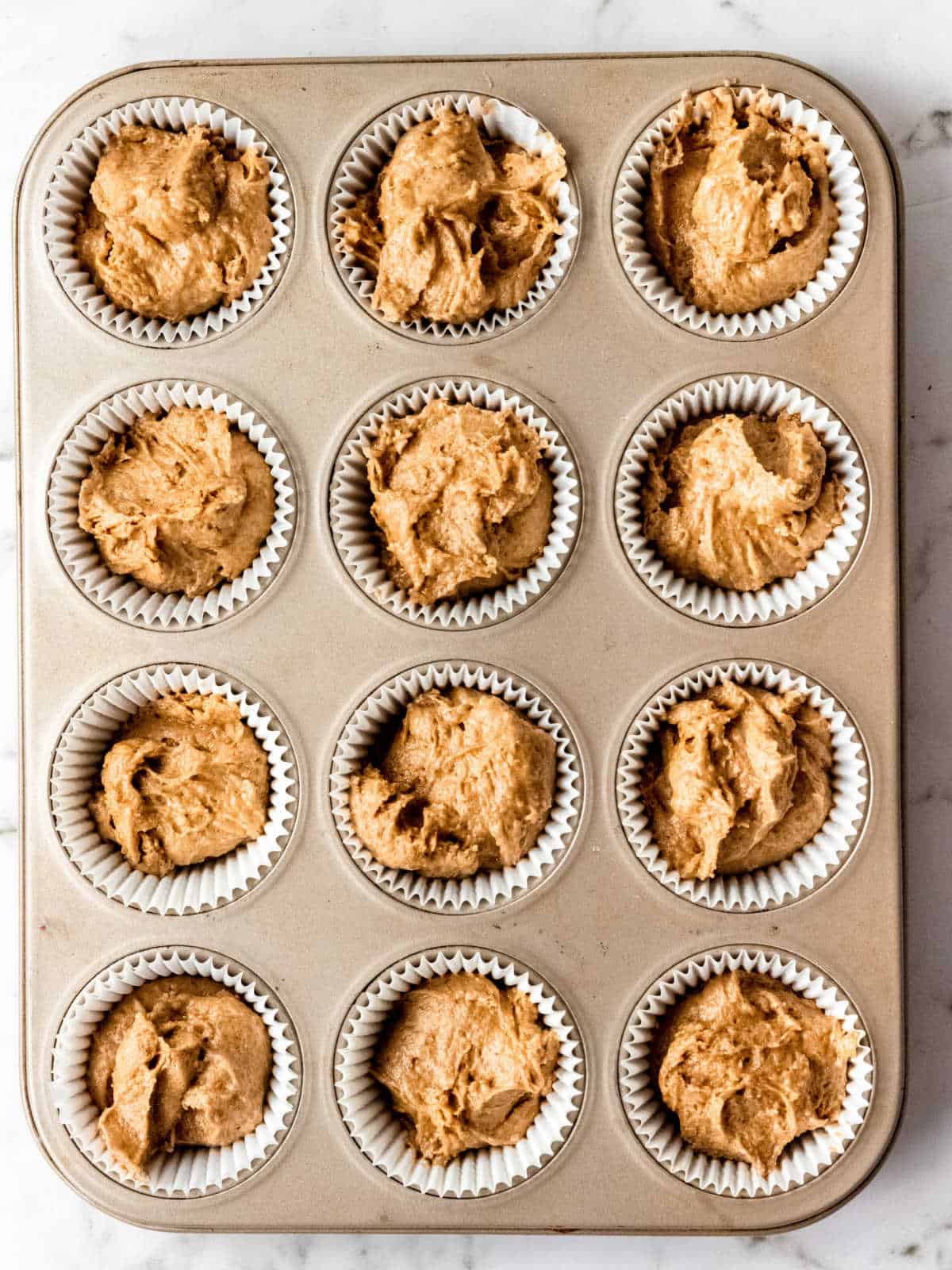 Muffin pan with white paper liners containing pumpkin muffin batter. Top view.