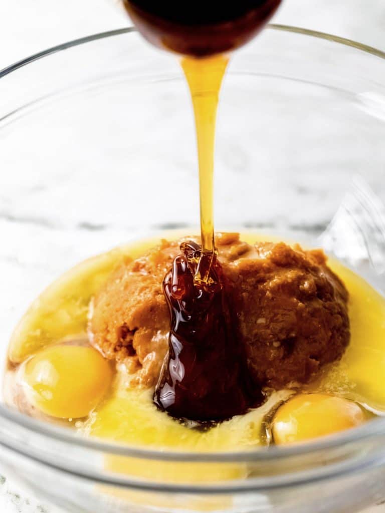 Glass bowl with eggs, maple syrup, melted butter, and brown sugar. White surface.