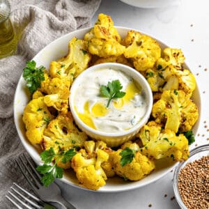 Round white platter of roasted cauliflower bites. White bowl with yogurt dip. Gray background.