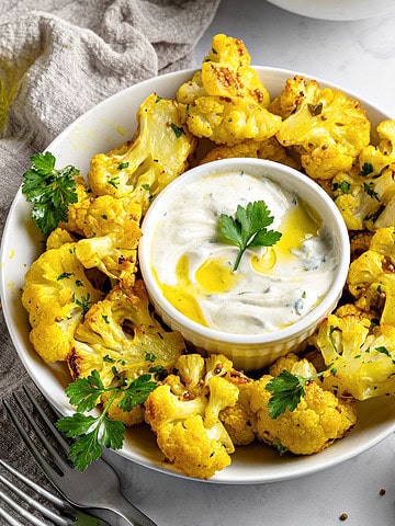 Round white platter of roasted cauliflower bites. White bowl with yogurt dip. Gray background.