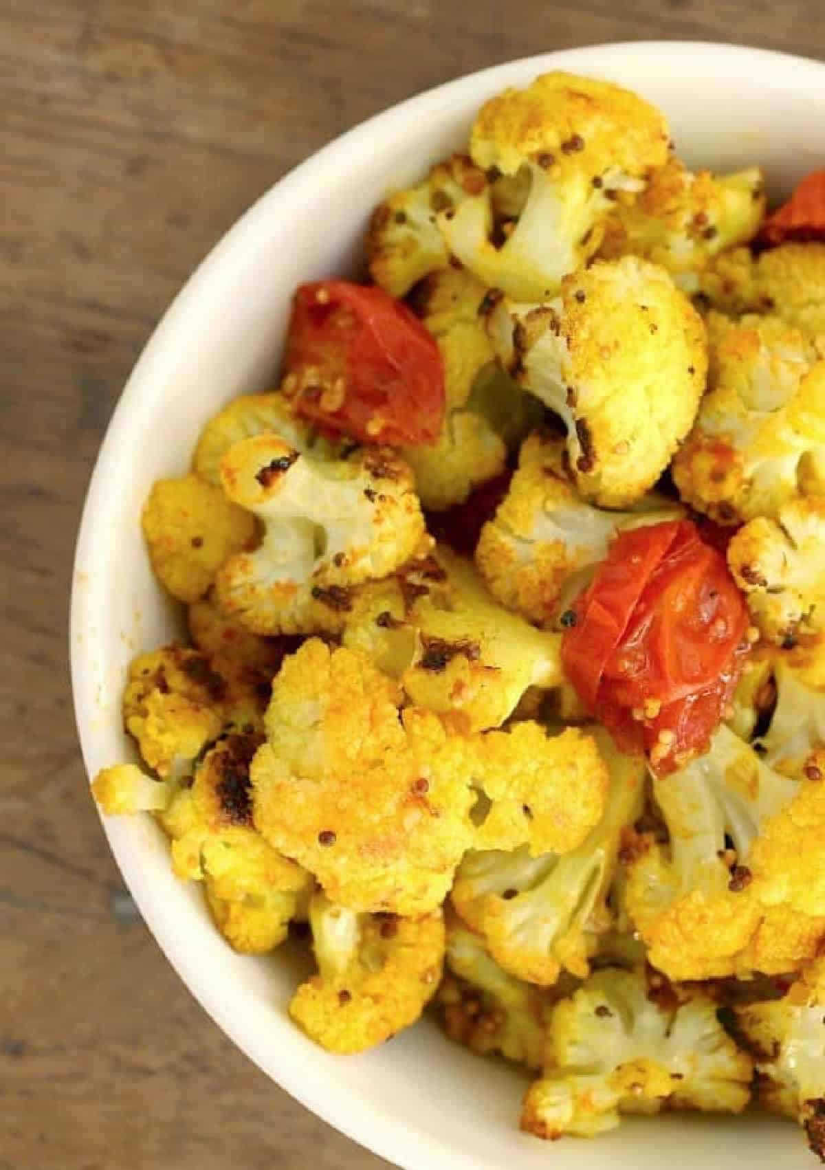 White bowl of yellow cauliflower pieces with tomatoes, on a wooden table. Partial view.