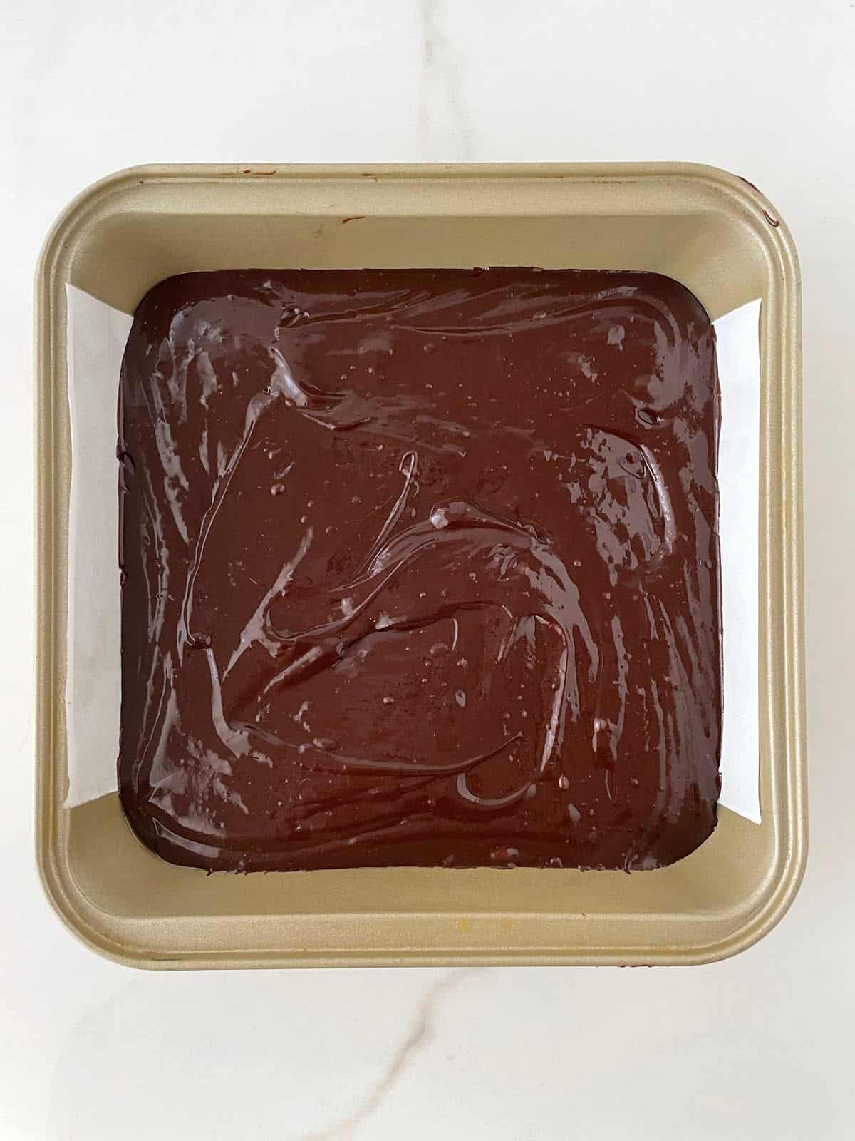 Square gold pan lined with parchment paper, with brownie batter on a white marble surface.