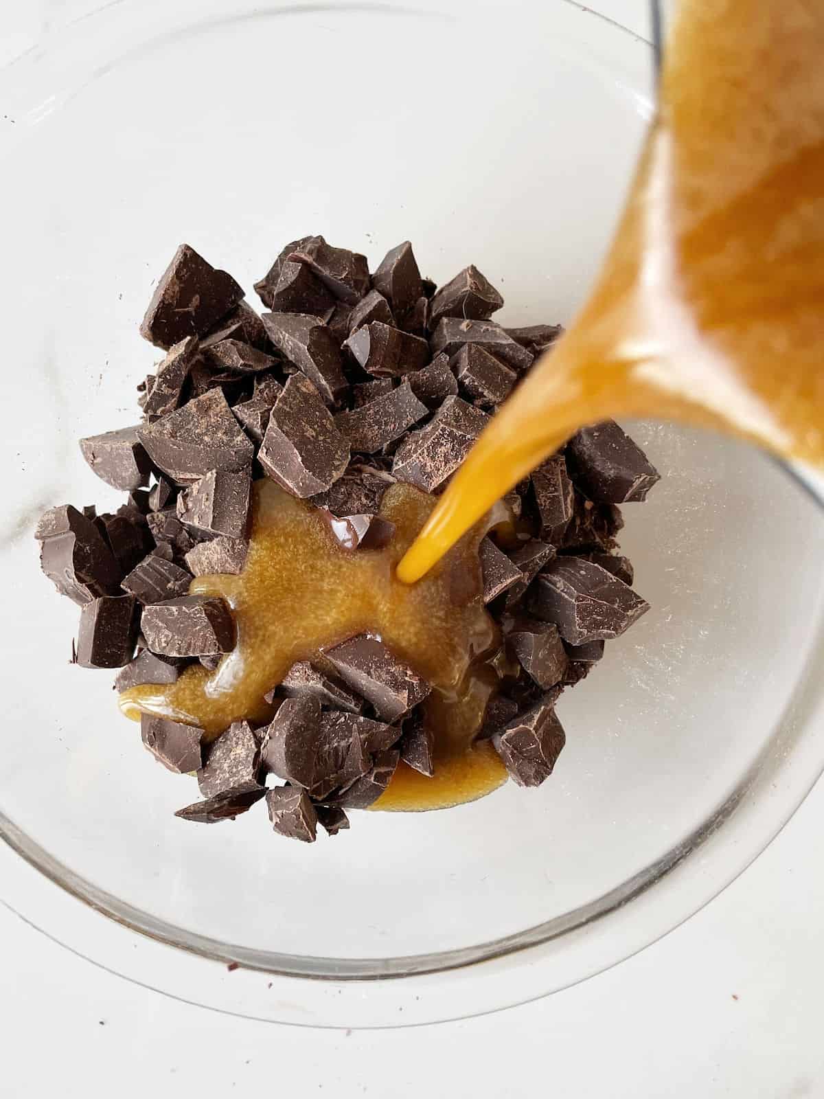 Melted brown butter mixture being poured over chopped chocolate in a glass bowl on a white surface. 