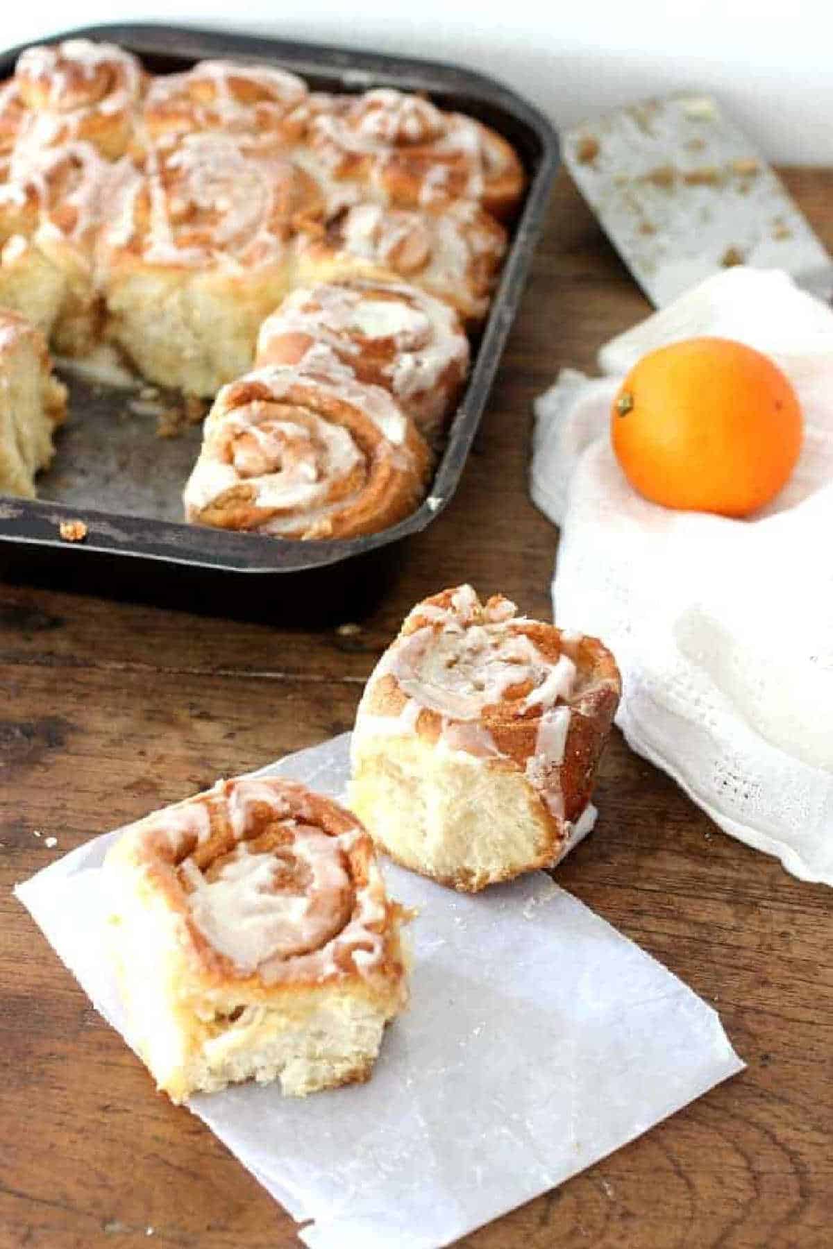 Wooden table with two cinnamon rolls, rest in metal pan, a white linen, an orange.
