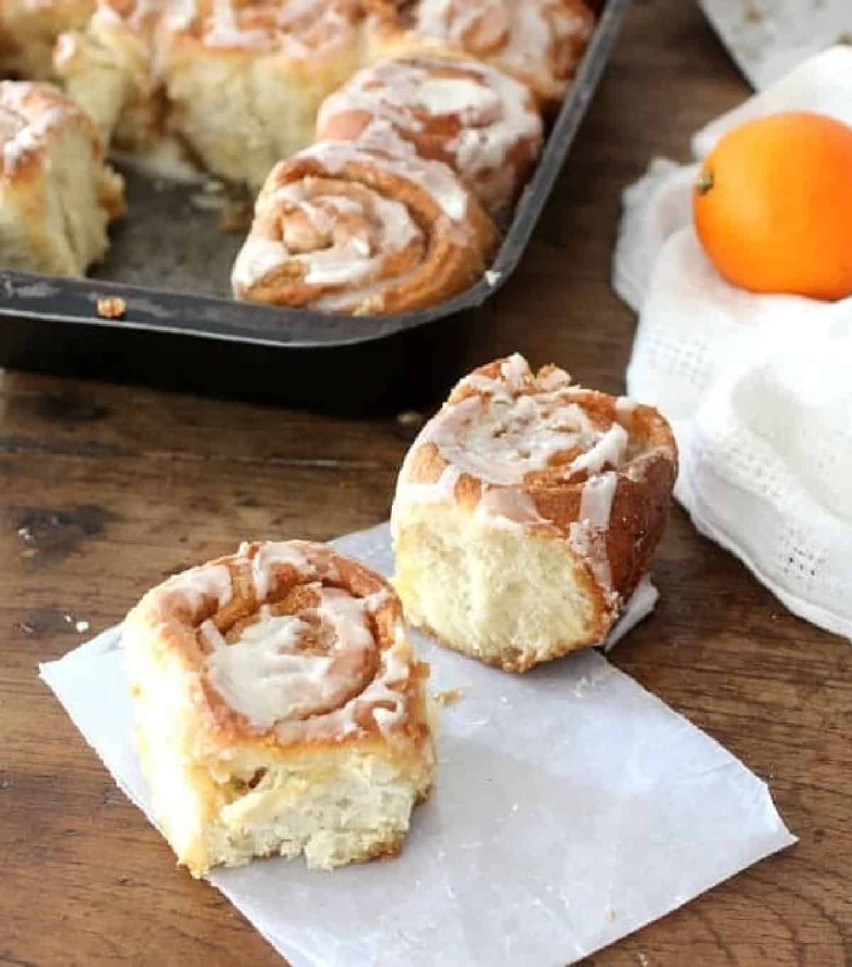 Two cinnamon rolls on wooden table, rest in pan, white linen and orange in background.