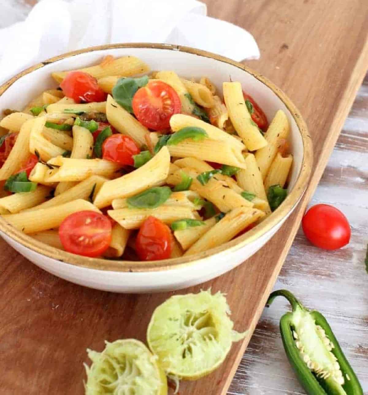 Bowl with short pasta and tomatoes on wooden board, limes and jalapeño around.