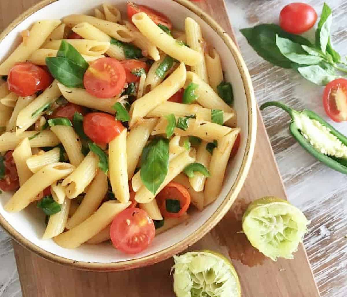 Top view of short pasta and tomatoes in bowl, white table, lime wedges, jalapeño and basil.