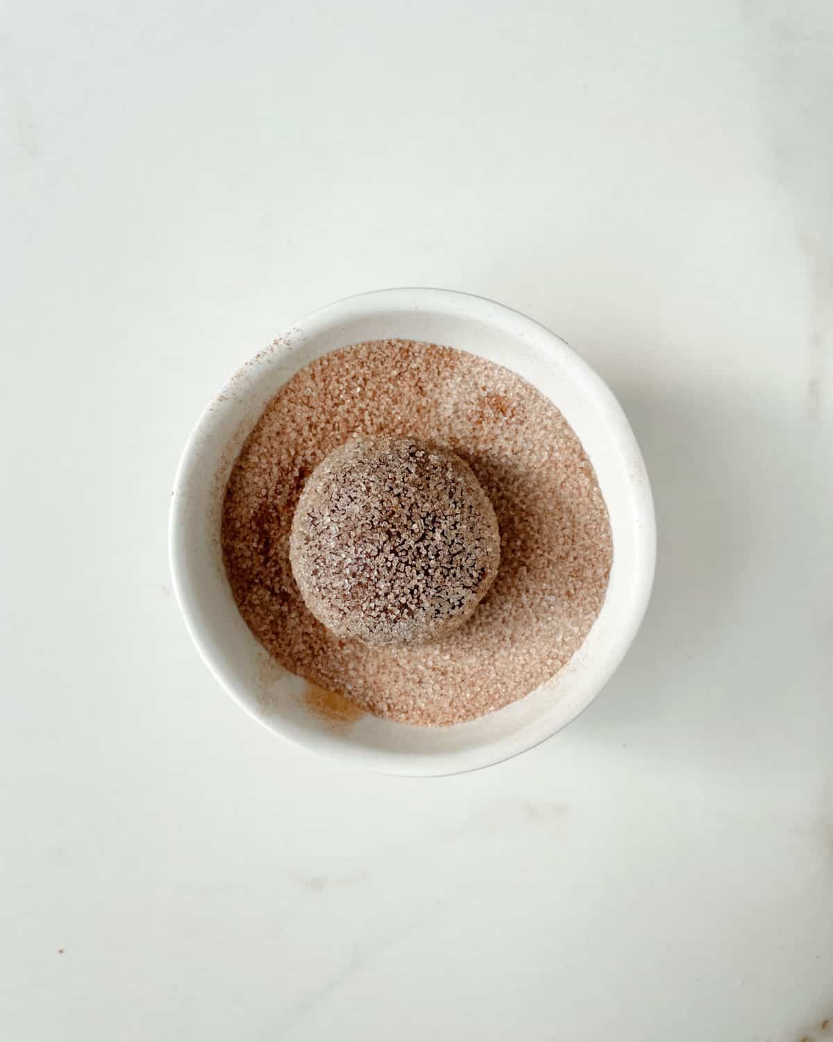 Cinnamon sugar in a white bowl with a cookie ball inside. White marble surface. 