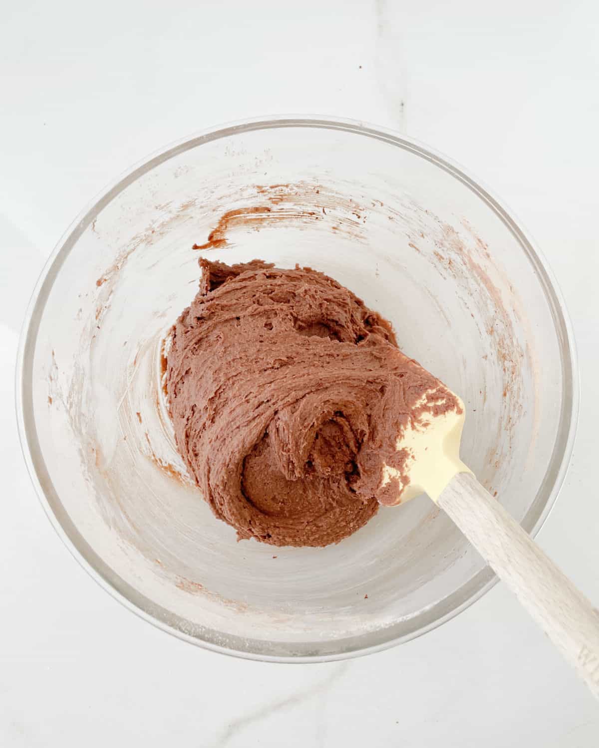 Chocolate cookie dough on a glass bowl with a spatula. White marble surface. 