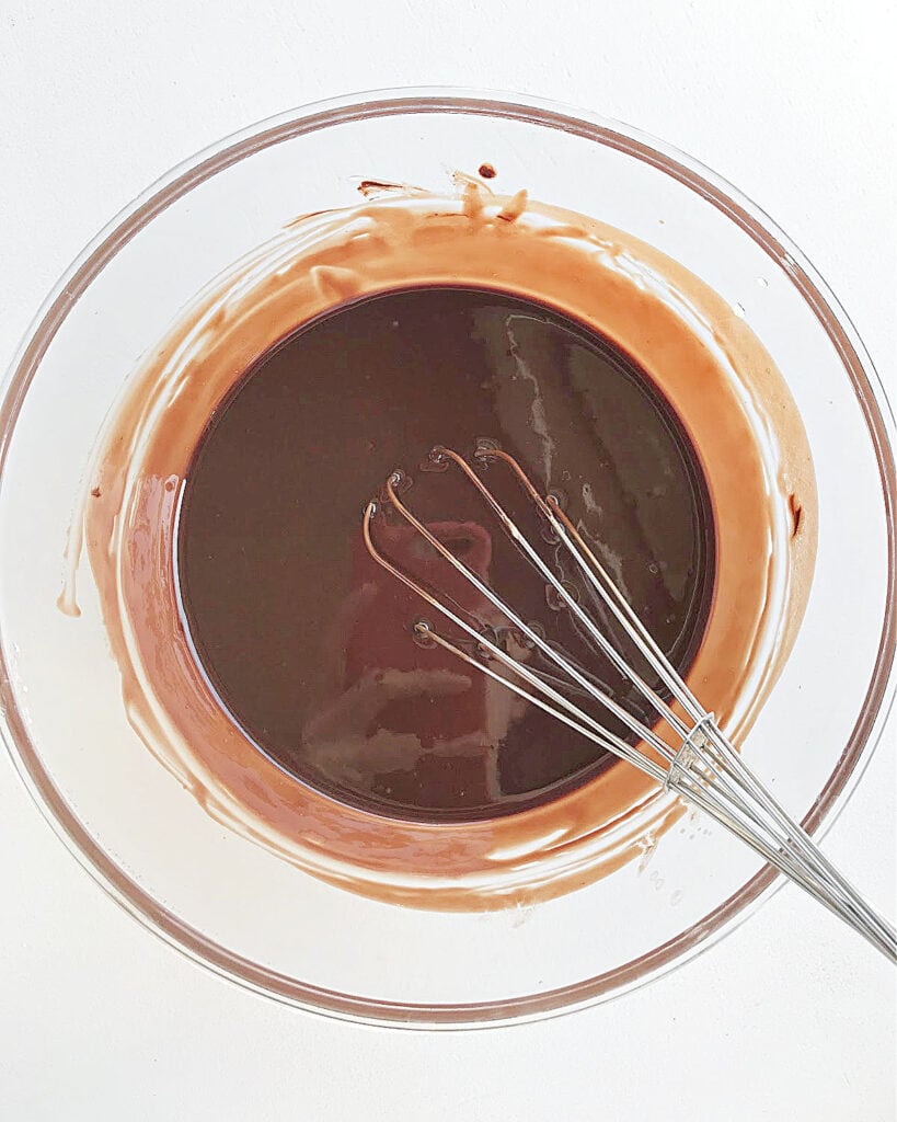 Glass bowl with melted chocolate and butter. A wire whisk. White background.