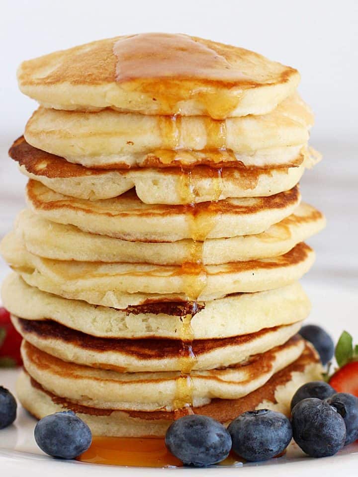 Tall stack of buttermilk pancakes with maple syrup and berries around. White background.
