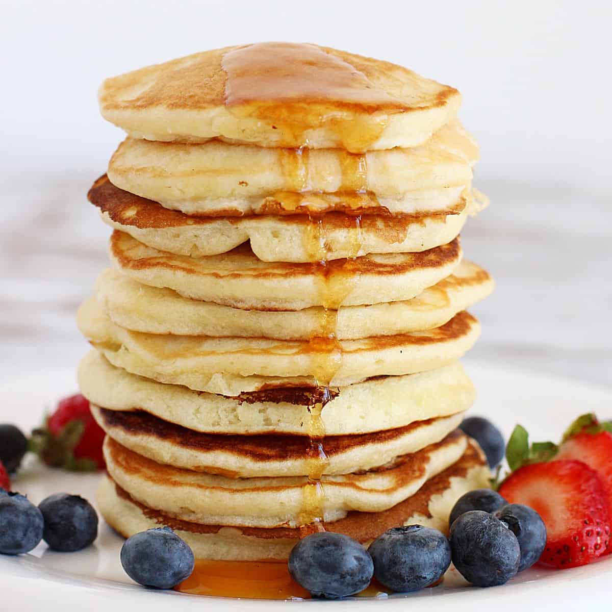 Tall stack of buttermilk pancakes with maple syrup and berries around. White background.