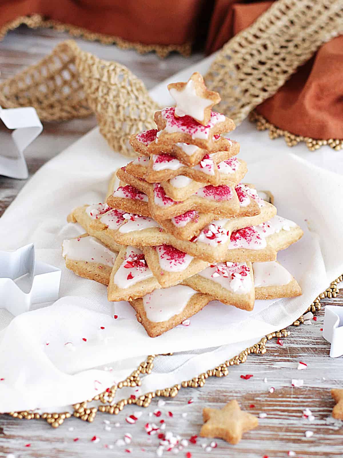 Star shaped Christmas tree made with frosted sugar cookie, candy cane crushed around, white and gold props, surface, and linens.