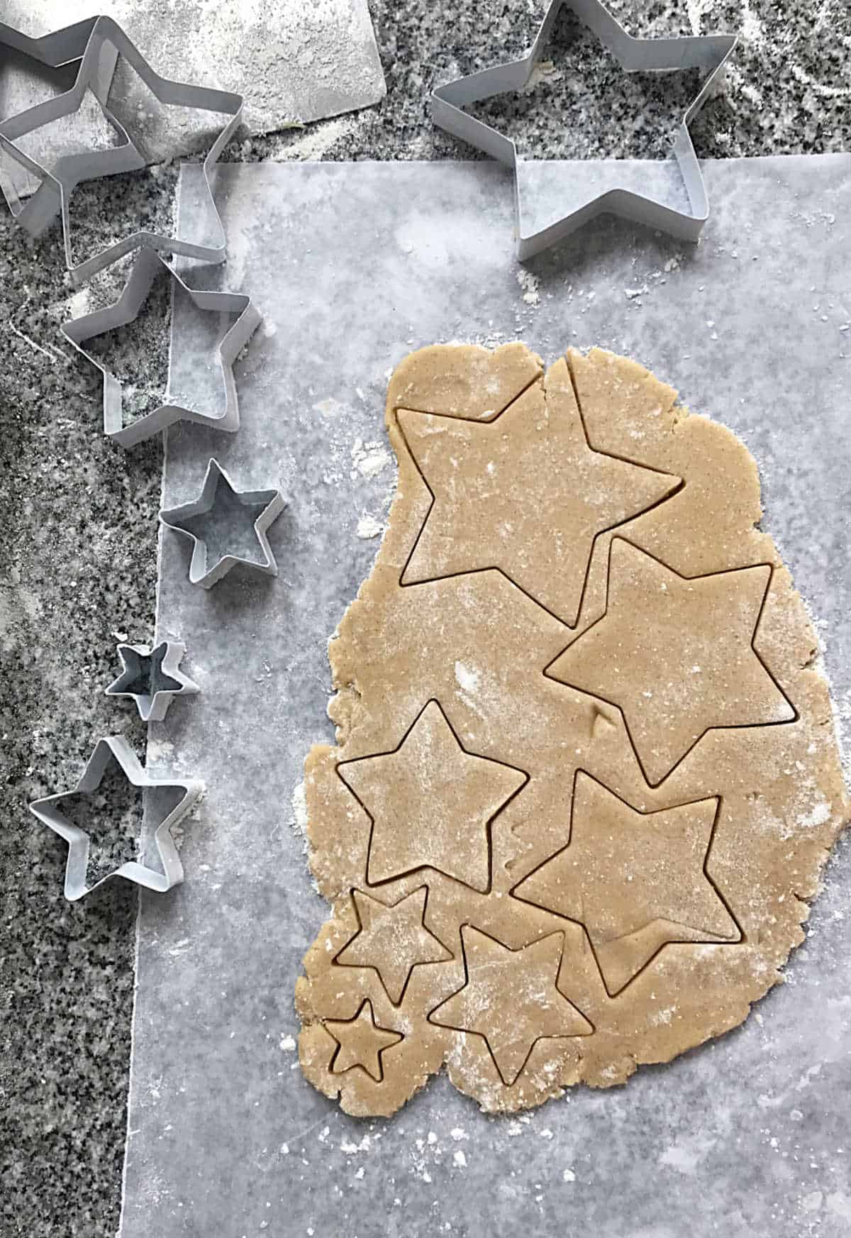Piece of parchment paper with cookie cut outs on grey counter, metal star shaped cutters around