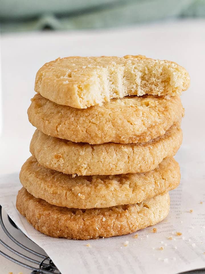 Close up of bitten coconut shortbread butter cookie on top of a stack. White parchment paper and surface.