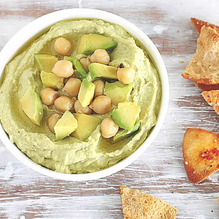 Washed white table with bowl containing avocado dip, chickpeas on top, pita chips around